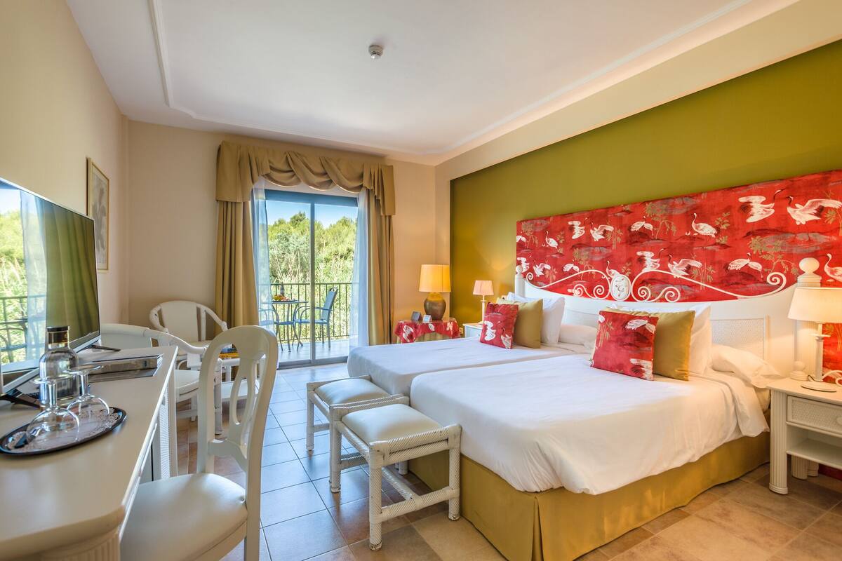 Habitación de hotel con dos camas, un escritorio con computadora, sillas y un balcón con vista a árboles.