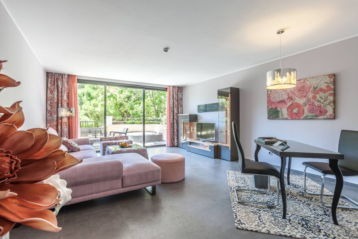 Una sala moderna con sofá, reposapiés y una mesa de comedor con sillas. Cuenta con un ventanal con cortinas, un televisor de pantalla plana y una planta decorativa.