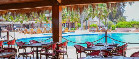 Un restaurante junto a la piscina con terraza, piscina y jardines tropicales.