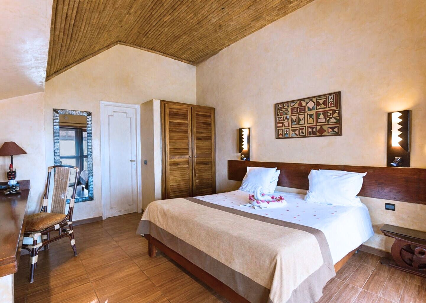 A bedroom with a large bed, wooden wardrobe, a chair, a desk, and a decorative wall piece.