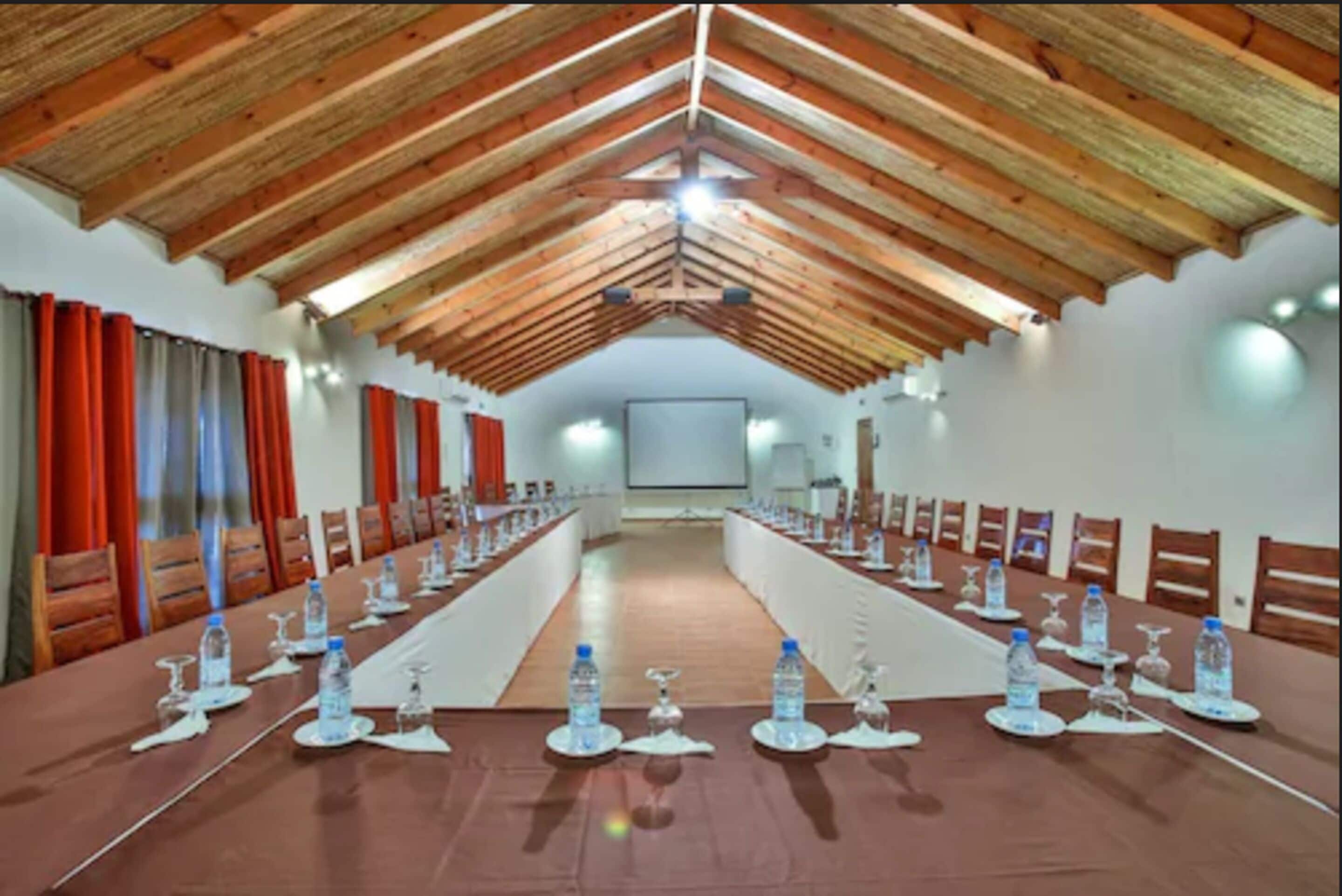 A conference room with a long table set for meetings, wooden ceiling beams, and red curtains.