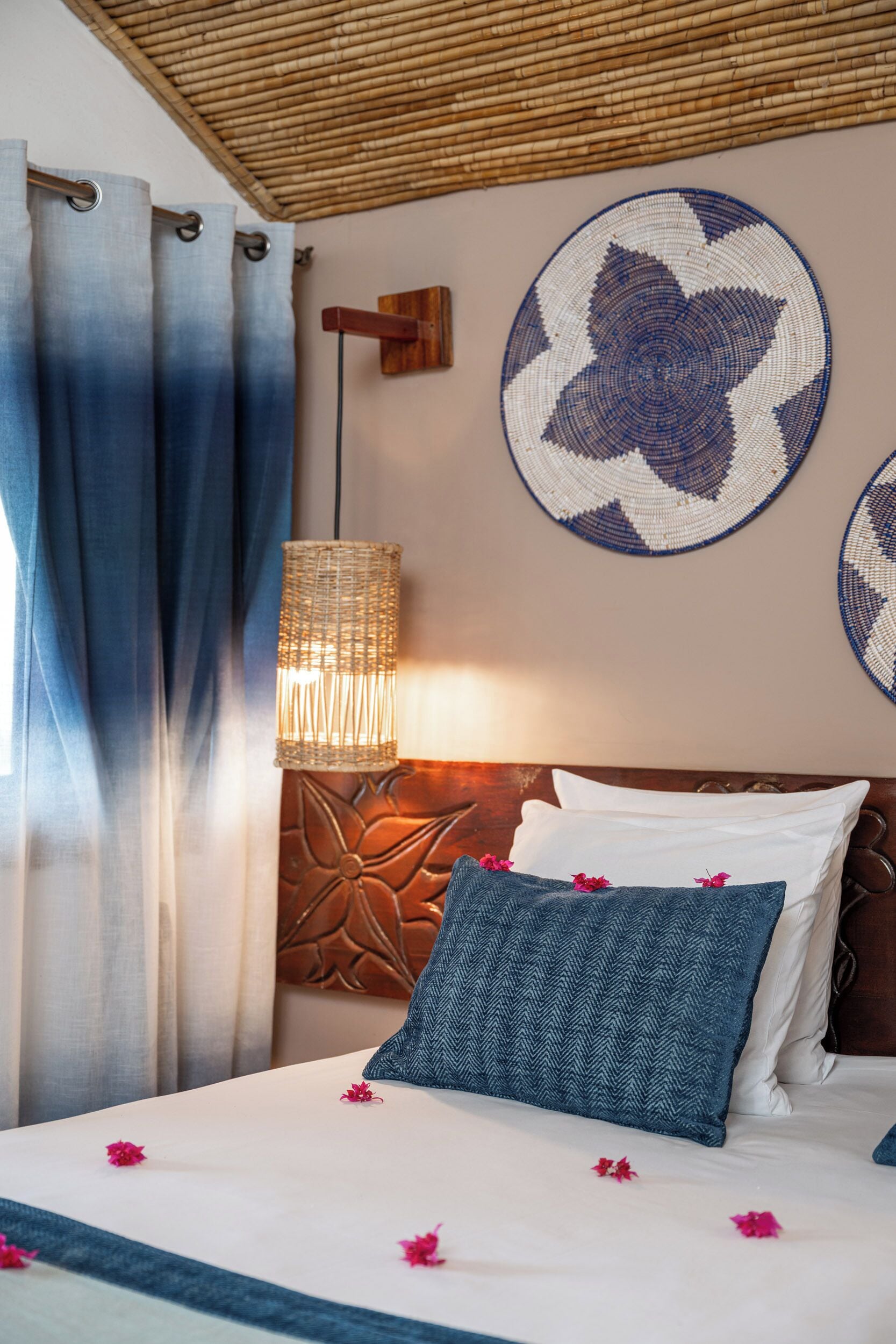 A bed with a white and blue patterned pillow, a woven lamp, and a wall with circular woven art.