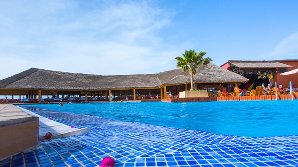 A swimming pool with a tiled deck, palm trees, and thatched-roof structures. - Royal Horizon Baobab - All inclusive (La Somone)