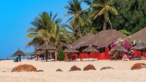 A beach with thatched-roof huts, palm trees, and a red building with a pink flower arrangement. - Royal Horizon Baobab - All inclusive (La Somone)