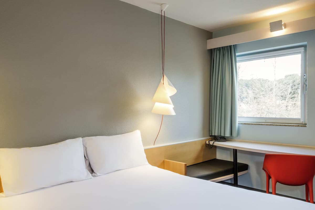 Une chambre d’hôtel avec un lit, un bureau, une chaise, une lampe et une fenêtre avec des rideaux.