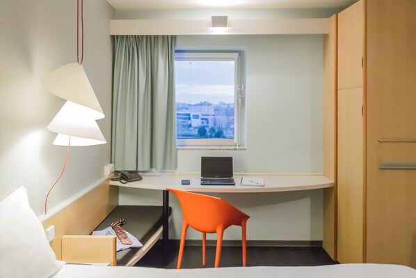 A hotel room with a desk, a chair, a bed, a window with a view of buildings, and a lamp.