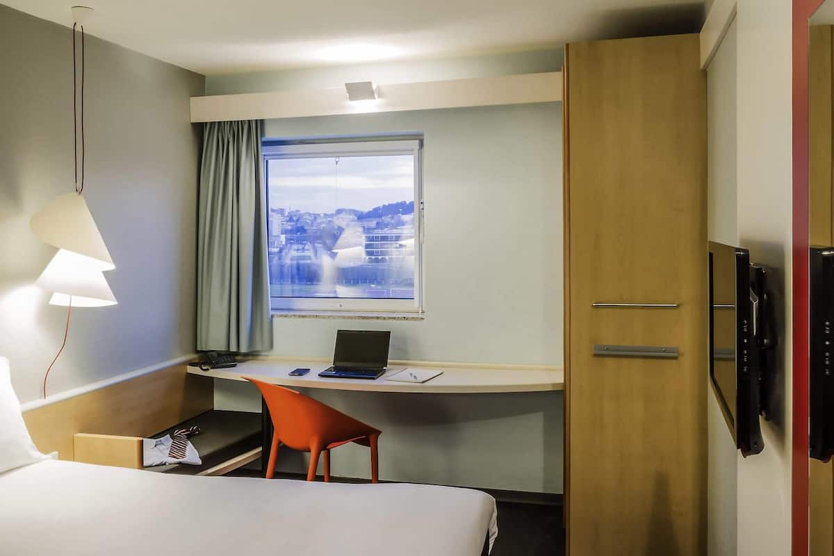 A hotel room with a bed, a desk with a laptop, a chair, a window with a view, and a TV.