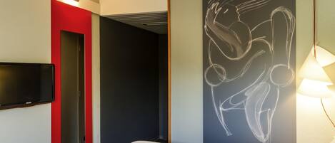 Una habitación de hotel con una cama, un televisor de pantalla plana en la pared, una puerta roja, una puerta negra y una pared con un dibujo en la pizarra.