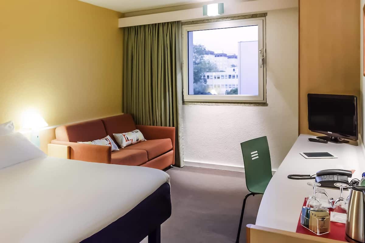 Une chambre d’hôtel comprenant un lit, un canapé, un bureau avec un ordinateur, une chaise et une fenêtre avec des rideaux.