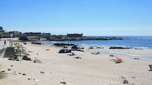 En strand med en stenmur, trappor som leder ner till stranden och en stenbrygga i bakgrunden.