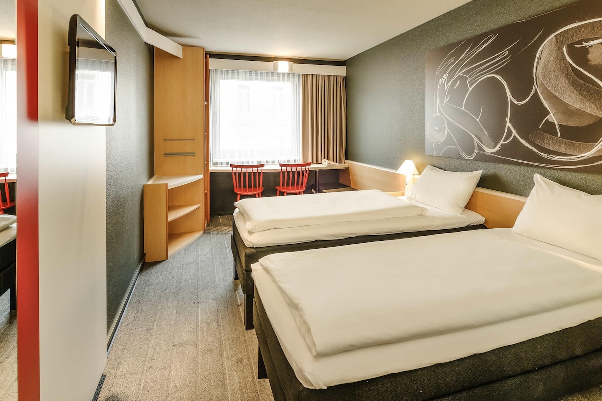 Une chambre d’hôtel avec deux lits, un bureau, deux chaises, une armoire et un grand tableau abstrait accroché au mur.