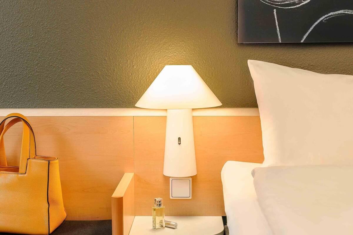 A hotel room with a wooden headboard, a white bedside table with a lamp, a bottle of perfume, and a yellow handbag.