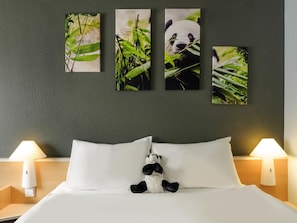 Uma cama com roupa de cama branca, um pelúcia de panda e quatro quadros emoldurados de pandas entre folhagem na parede.
