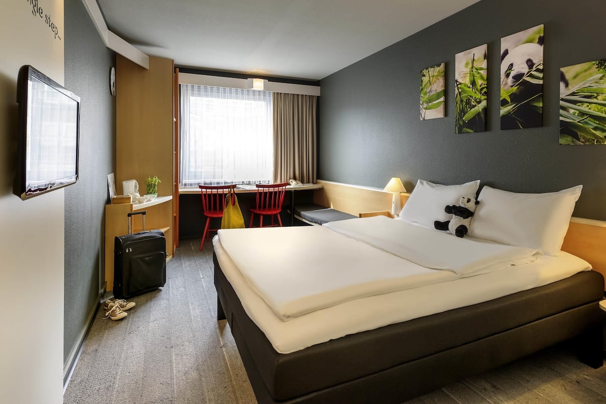 Une chambre d’hôtel avec un lit, un bureau, une chaise, une fenêtre avec des rideaux et des œuvres d’art accrochées au mur.