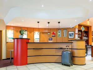 Zona de recepción de un hotel con mostrador de madera, un pilar rojo, una planta en maceta, un panel informativo digital y una zona de bar con vasos y botellas.
