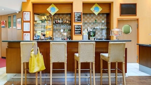 A bar area with wicker chairs, a counter with a cash register, and a selection of bottles and glasses. - ibis Wien City (Vienna)