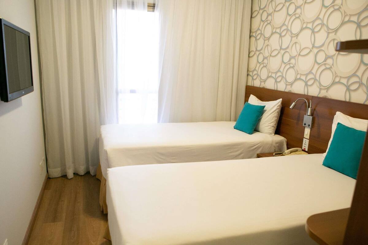 Habitació d'hotel amb dos llits, una televisió penjada a la paret i una finestra amb cortines.