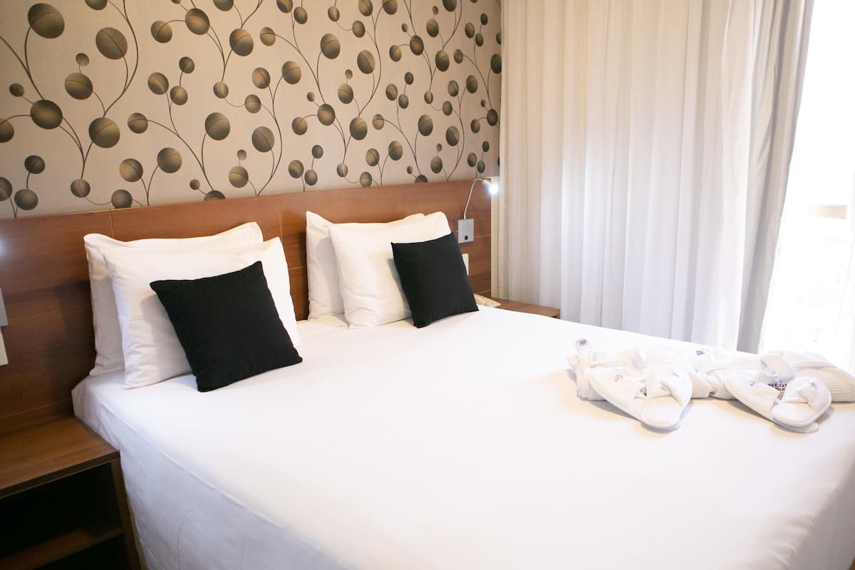 Una habitación de hotel con cama, almohadas, cabecera, mesita de noche y ventana con cortinas.