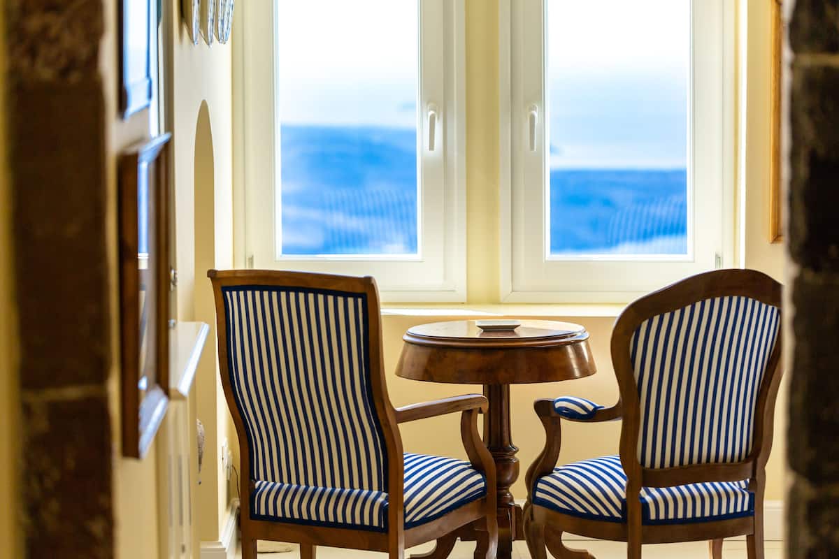 A room with two striped chairs and a round wooden table, a window with a view of the ocean, and a stone wall.