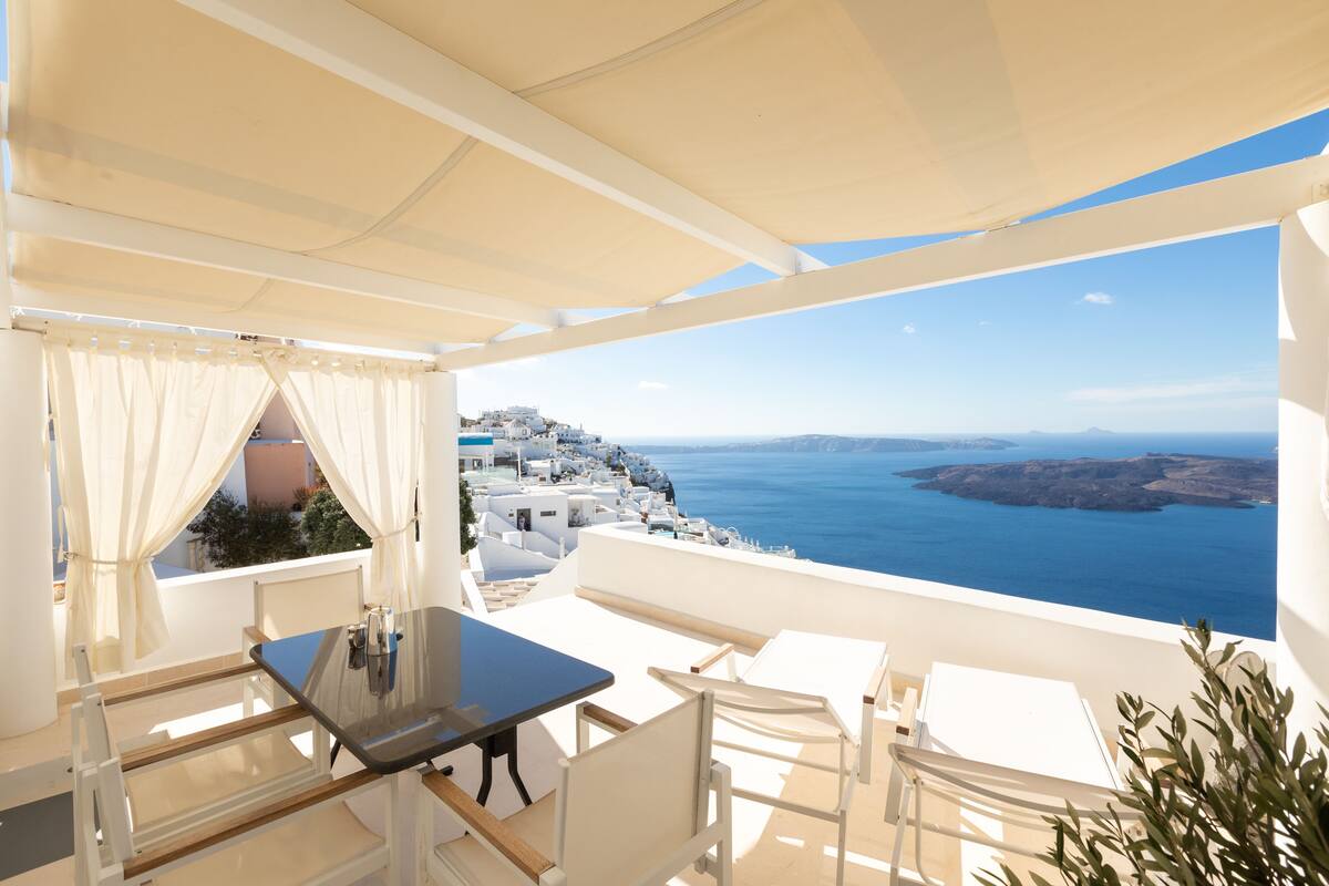 A terrace with a table and chairs, overlooking a coastal view with white buildings and the sea.