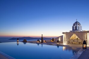 A infinity pool with a view of the sea and a building with a dome and white curtains. - The Tsitouras Collection Hotel (Santorini)