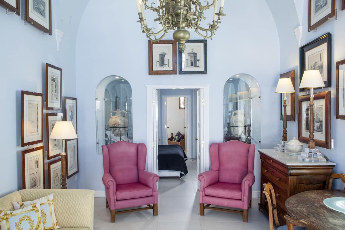 A room with two pink armchairs, a wooden dining table, and a chandelier. There are framed pictures on the walls and a bed with a blue bedspread in the background.