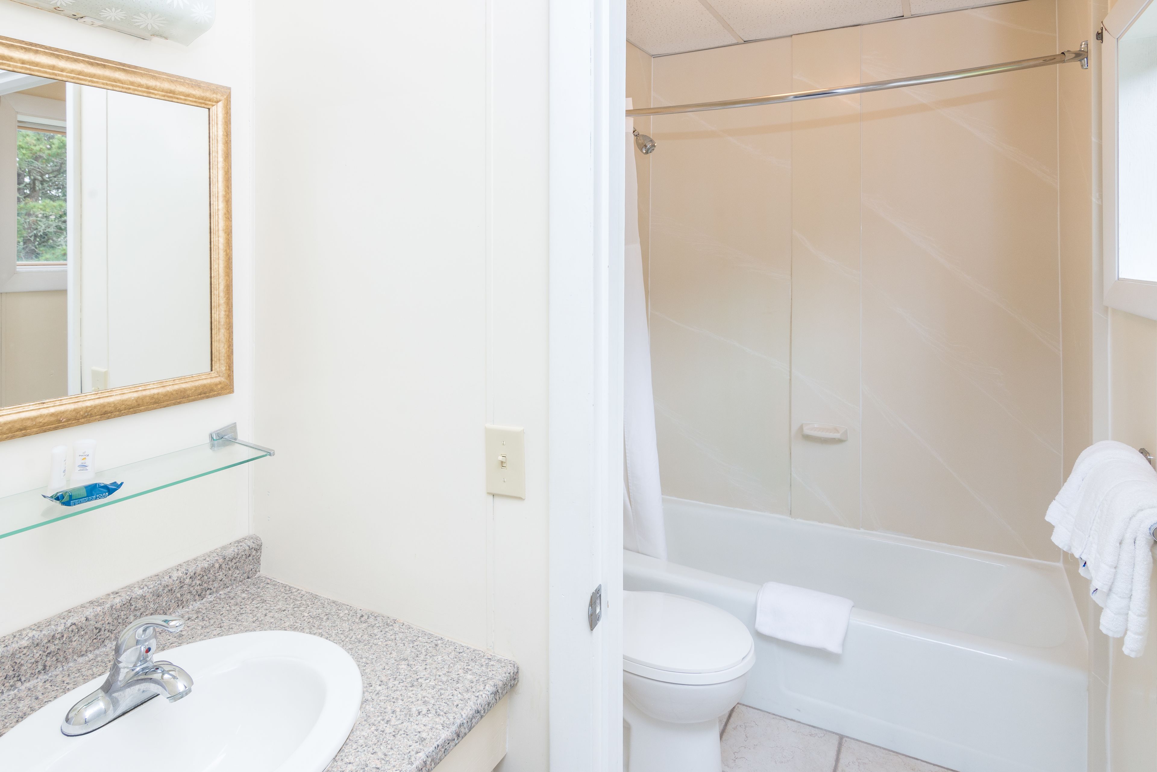 A bathroom with a granite countertop sink, a mirror with a gold frame, a glass shelf, a white toilet, and a white bathtub with a shower curtain.
