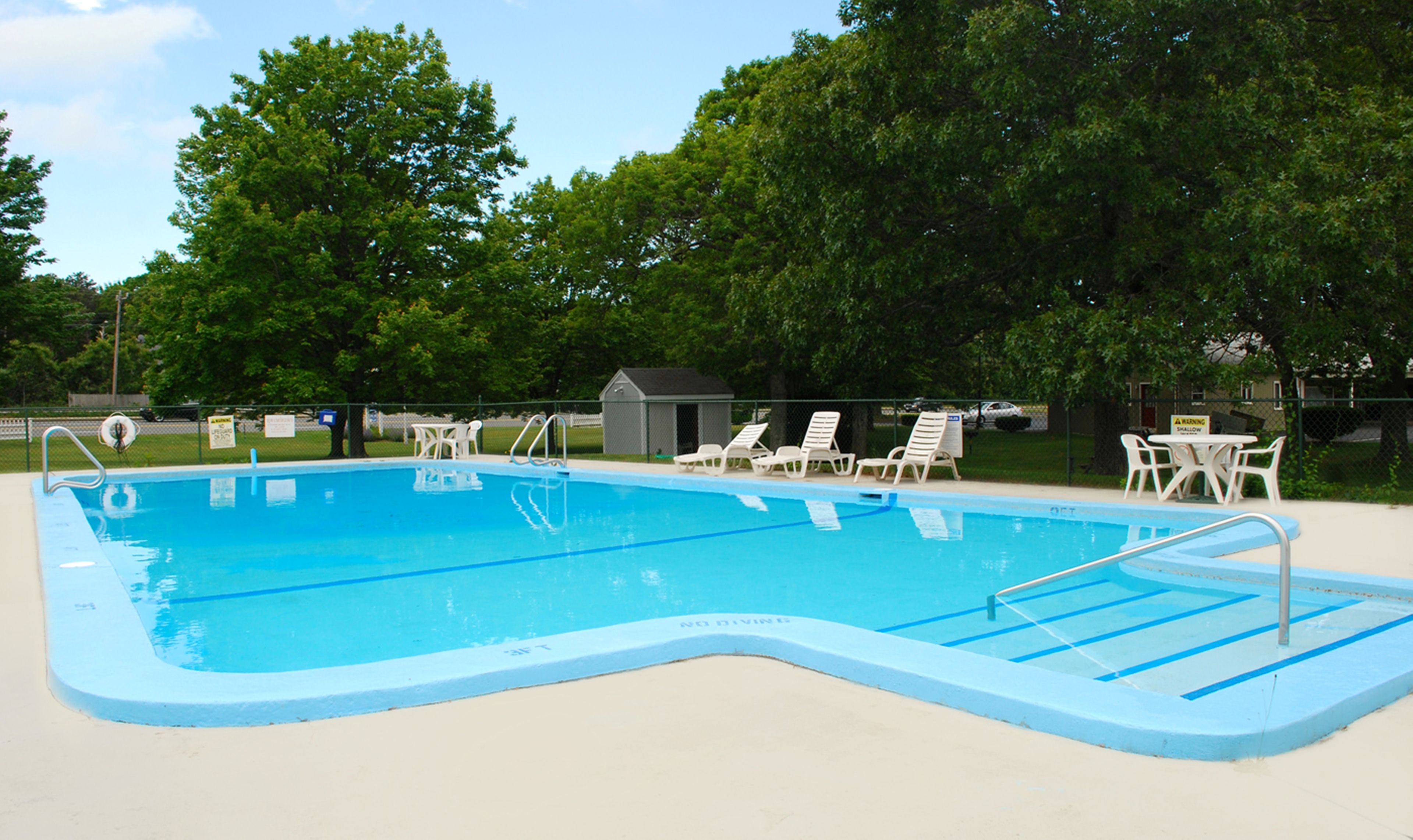 A rectangular swimming pool with a blue edge, surrounded by white lounge chairs and a small shed.