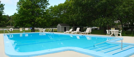 A rectangular swimming pool with a blue edge, surrounded by white lounge chairs and a small shed.