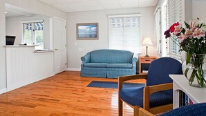 A room with a blue sofa, a wooden chair, a small table with a lamp, and a vase with flowers. - Ocean Park Inn (Eastham)