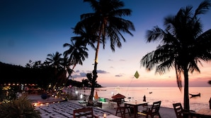 A beachside restaurant with outdoor seating, palm trees, and a view of the ocean at sunset. - Long Bay Resort (Ko Pha-ngan)