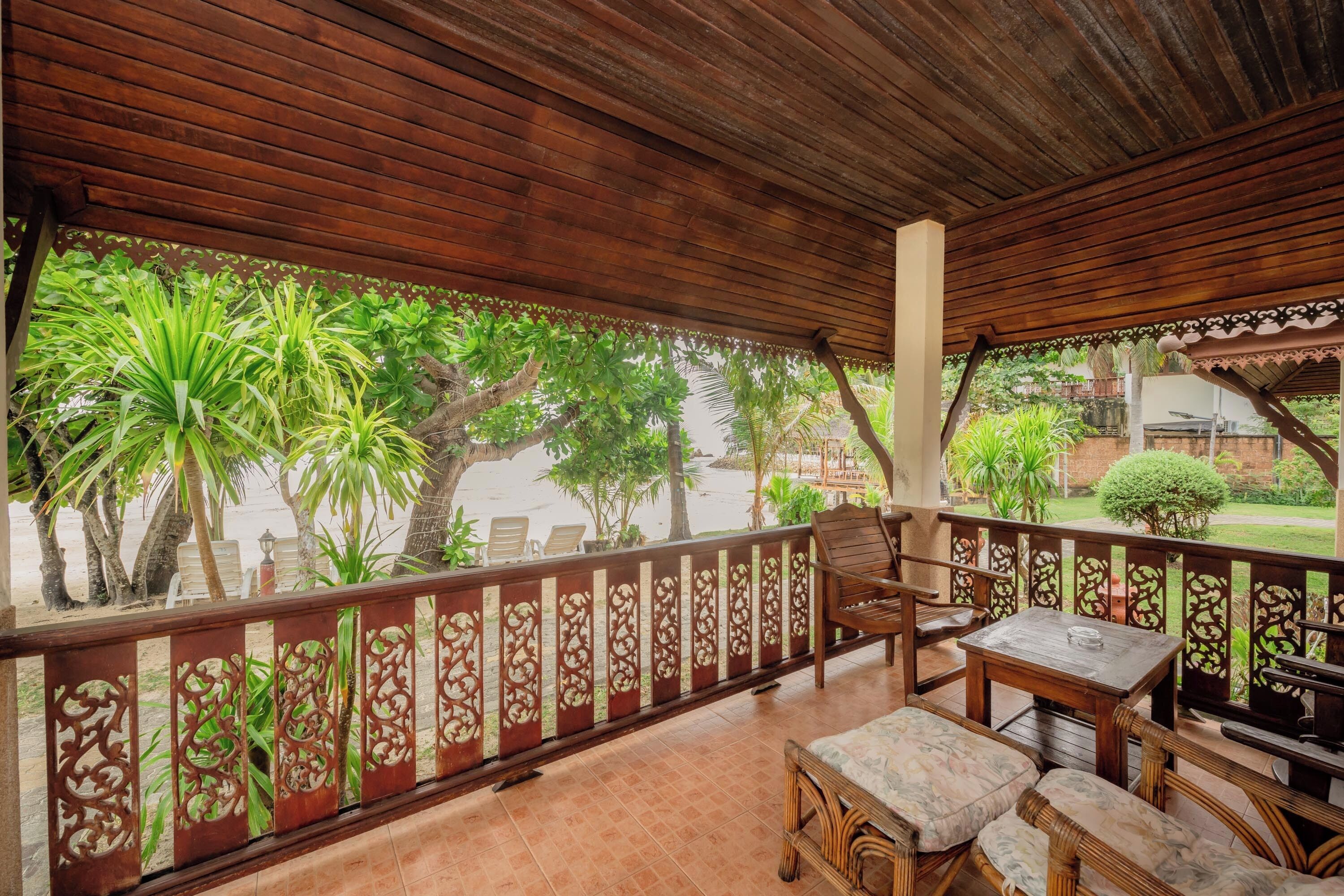 A covered outdoor seating area with a wooden table and chairs, surrounded by a decorative fence and lush greenery.