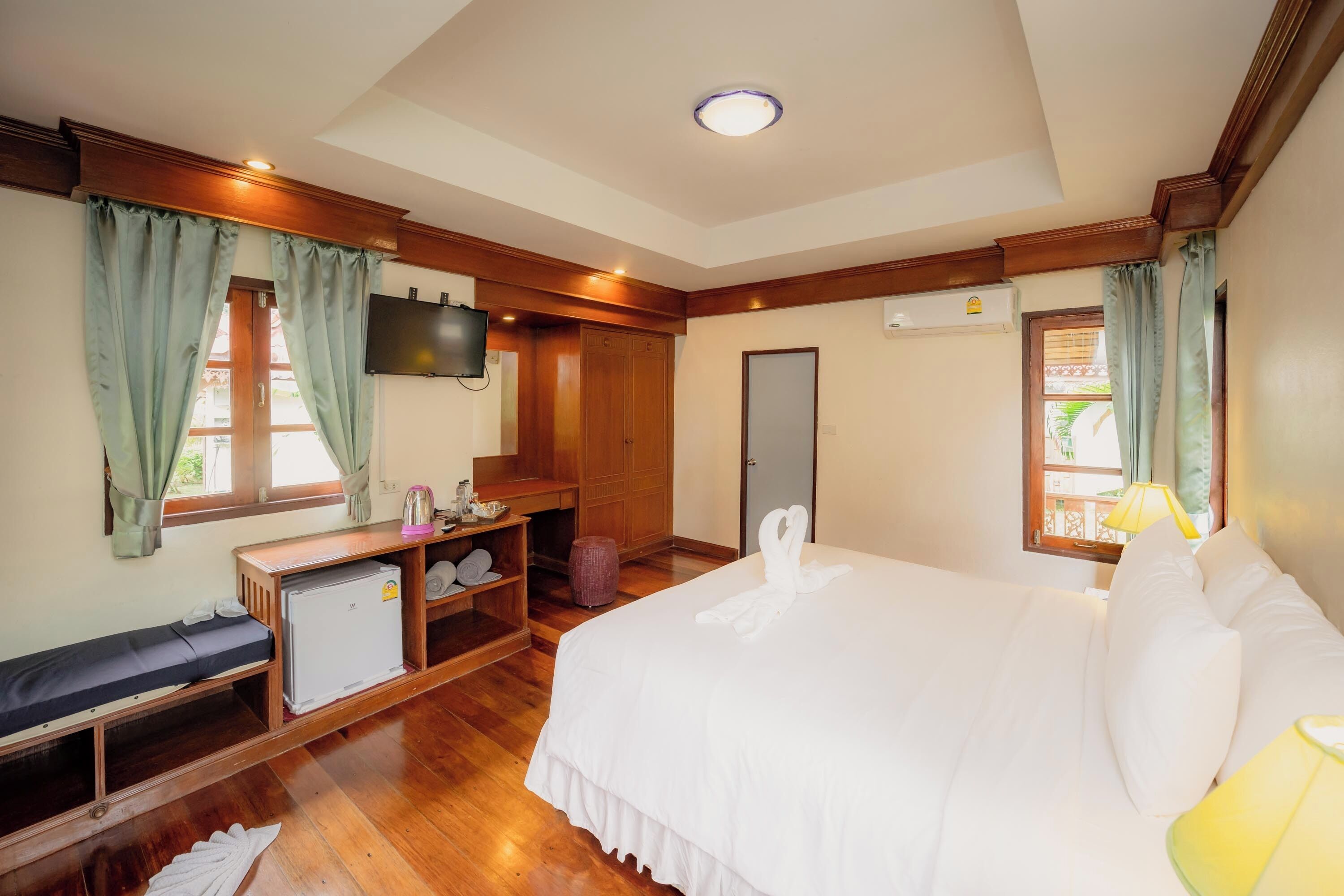 A hotel room with a large bed, a TV mounted on the wall, wooden furniture, and a window with curtains.
