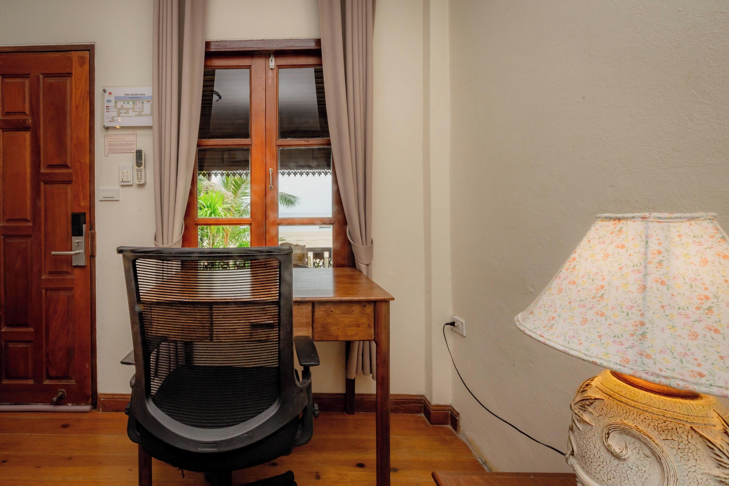 A room with a wooden door, a window with curtains, a table, a chair, and a lamp.