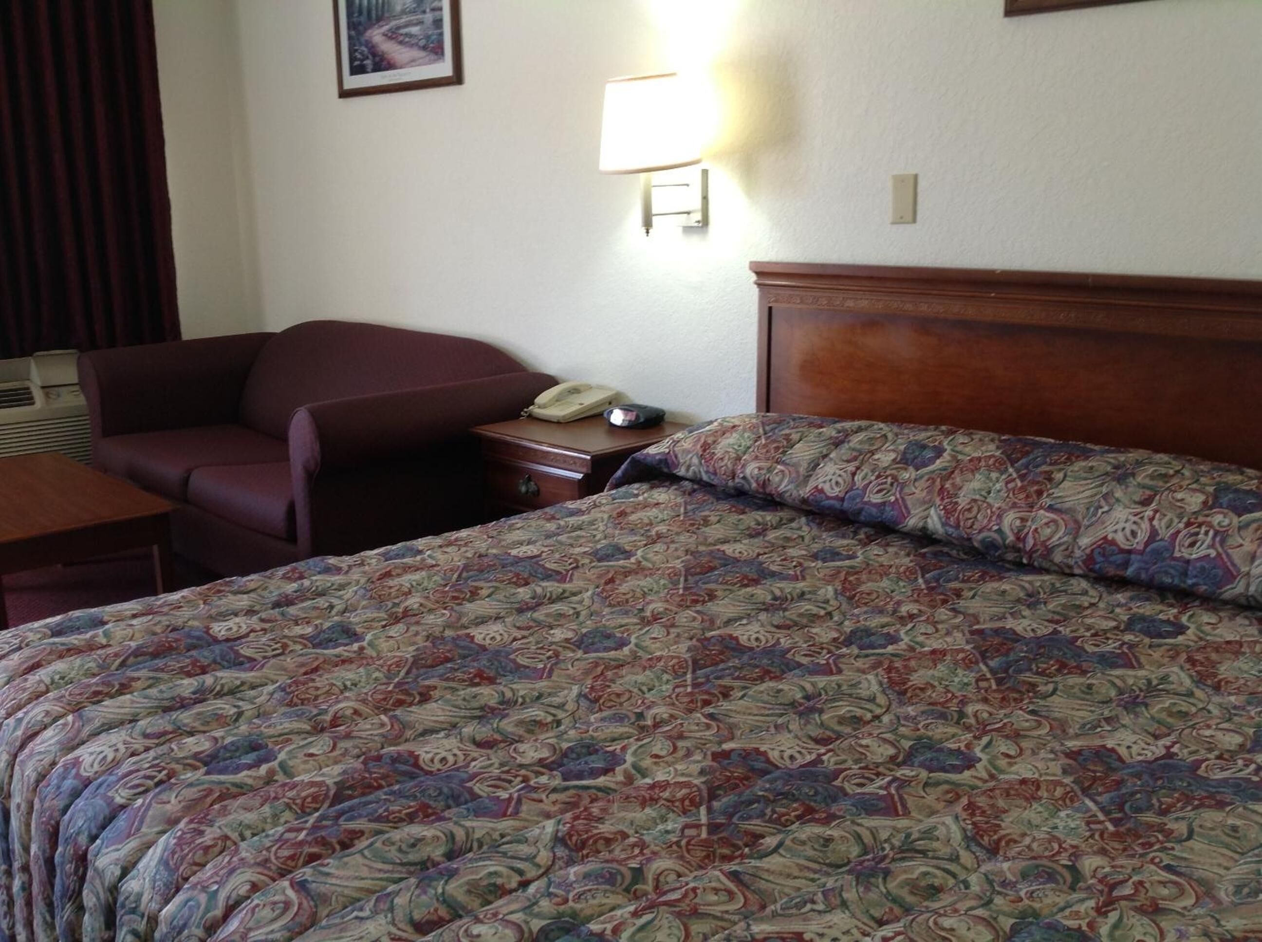 A hotel room with a large bed, a wooden headboard, a desk with a telephone, a chair, and a wall-mounted lamp.
