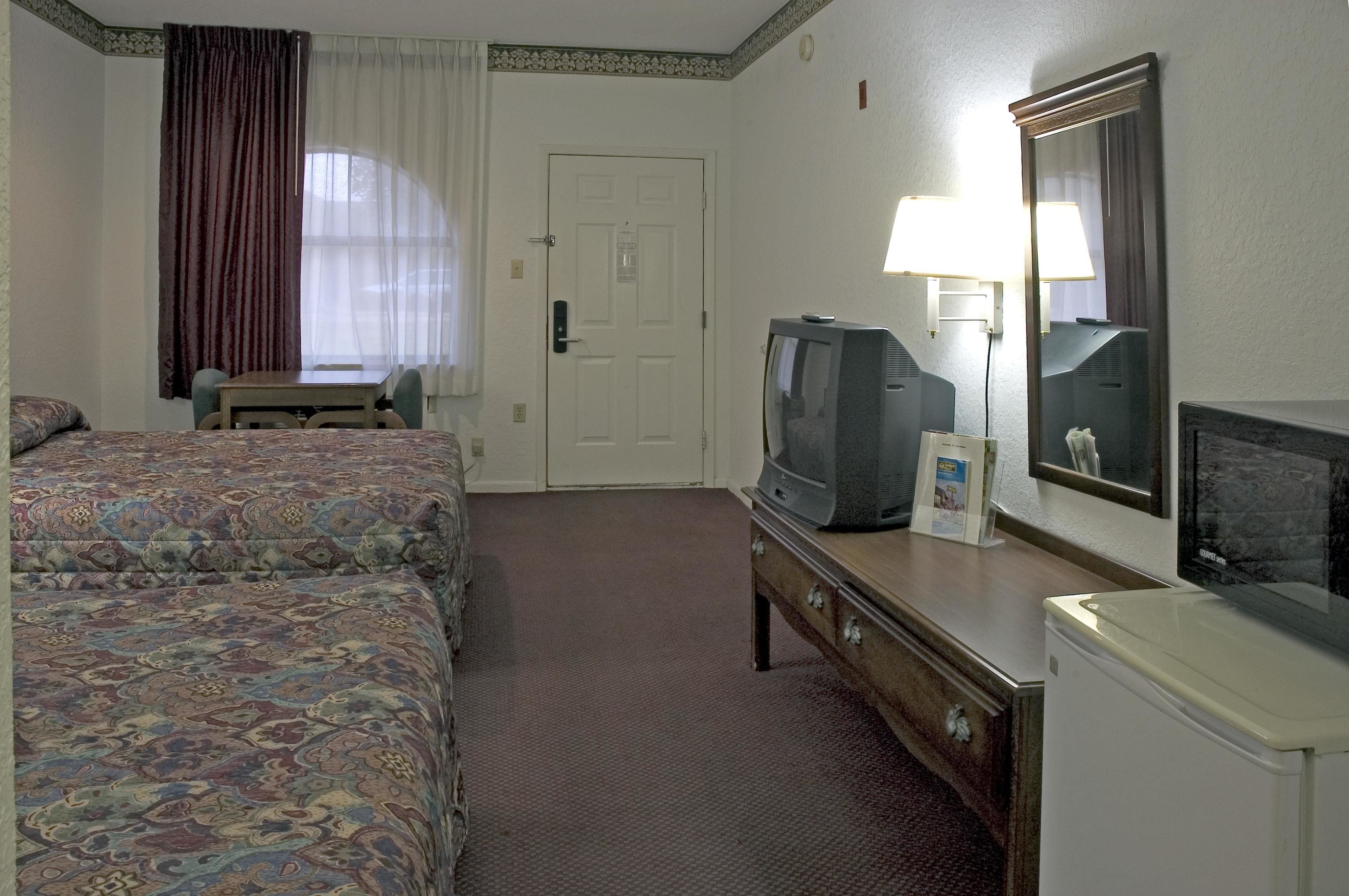 Habitación de hotel con dos camas, televisión, cómoda, espejo y ventana con cortinas.