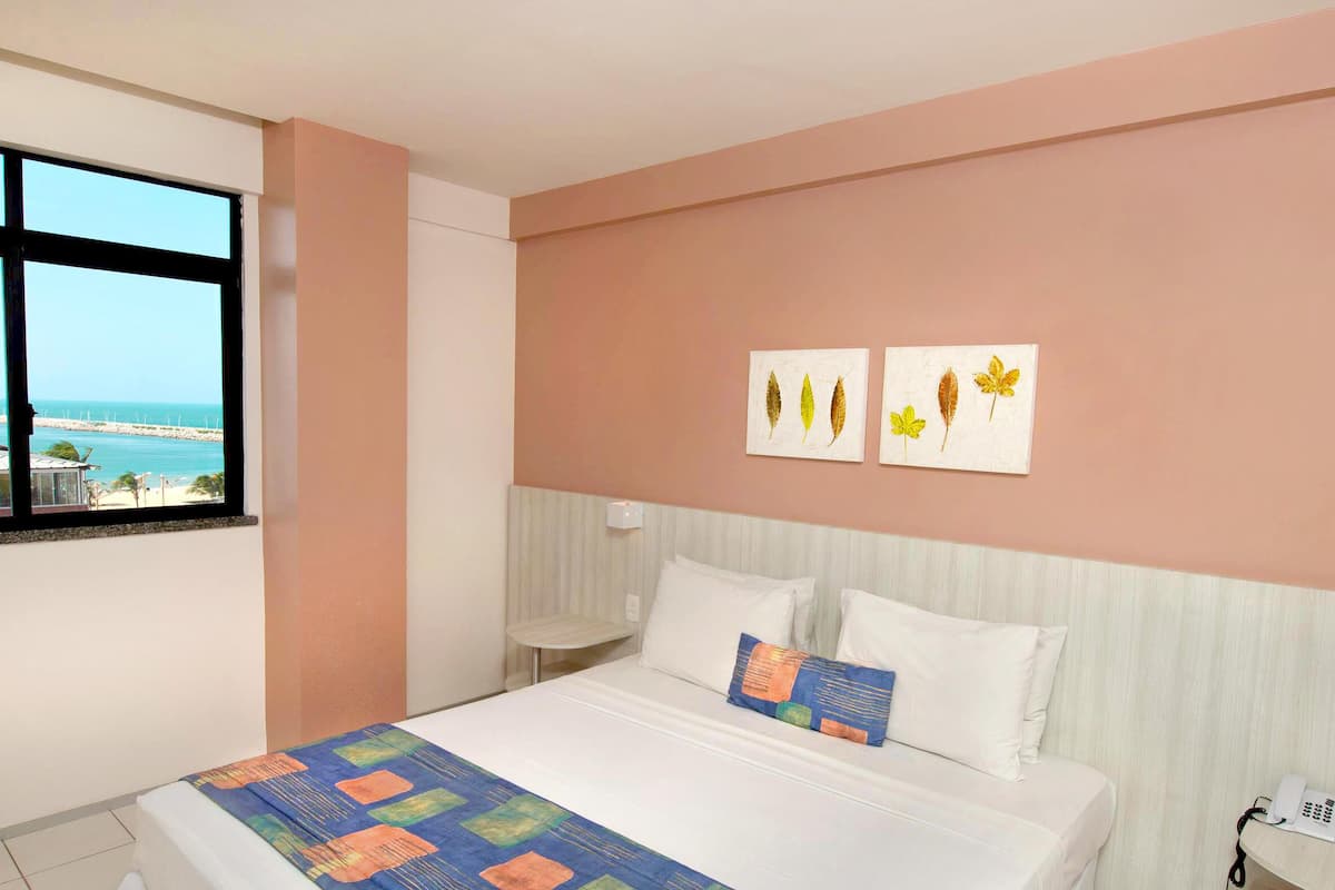 Habitación de hotel con cama, ventana con vista al mar y dos obras de arte en la pared.