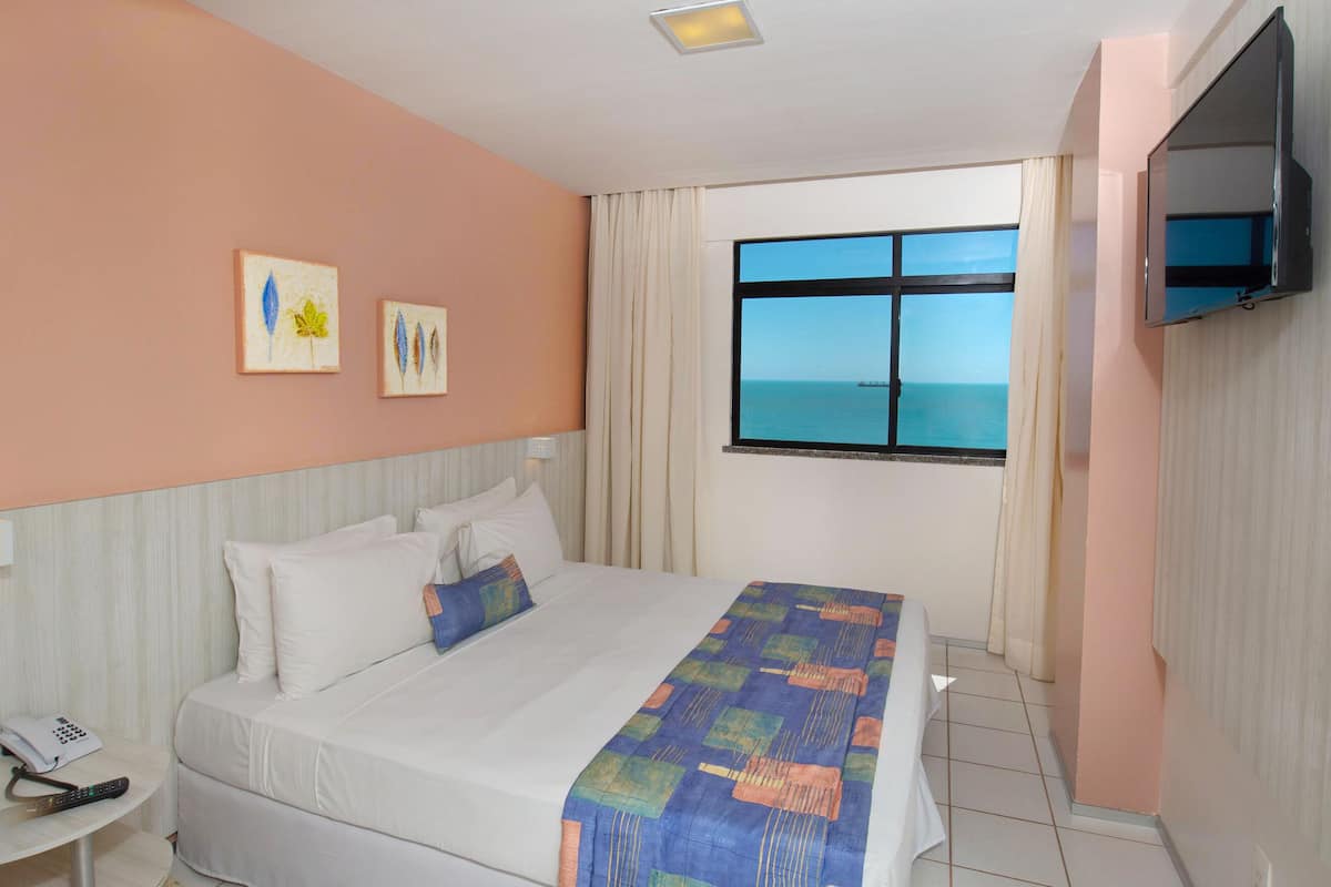 Habitación de hotel con cama, mesita de noche con teléfono, televisor montado en la pared y ventana con vista al mar.