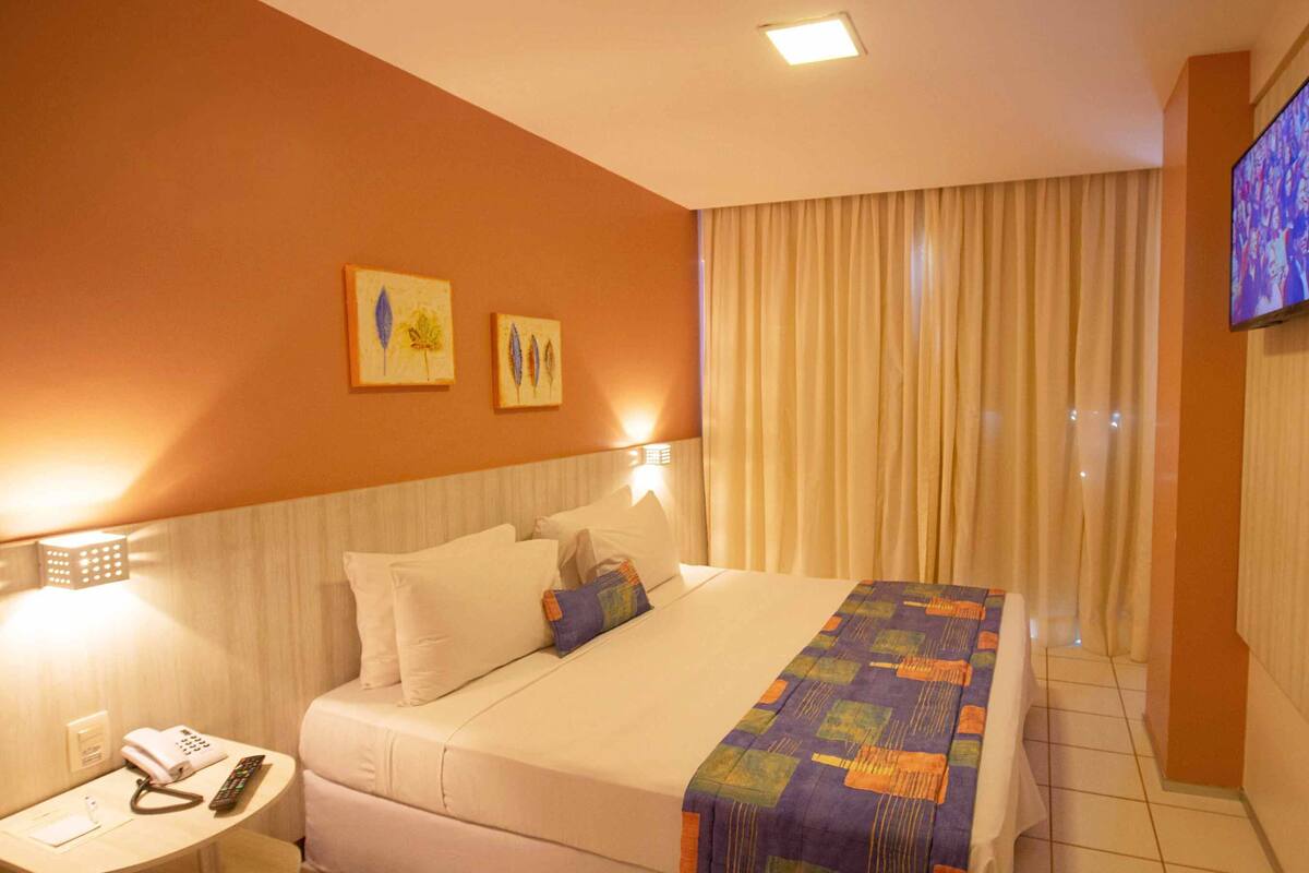 Una habitación de hotel con cama, televisión en la pared, mesita de noche con teléfono y control remoto, y ventana con cortinas.