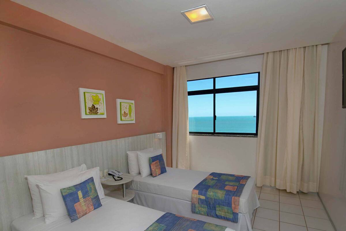 Habitación de hotel con dos camas, televisión, ventana con vista al mar y obras de arte en la pared.