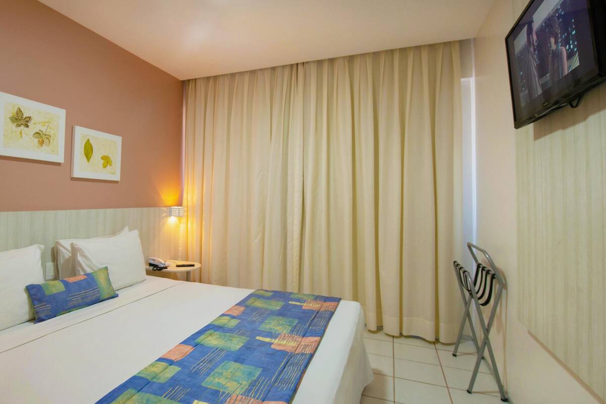Una habitación de hotel con cama, mesita de noche, lámpara, televisor y una silla.