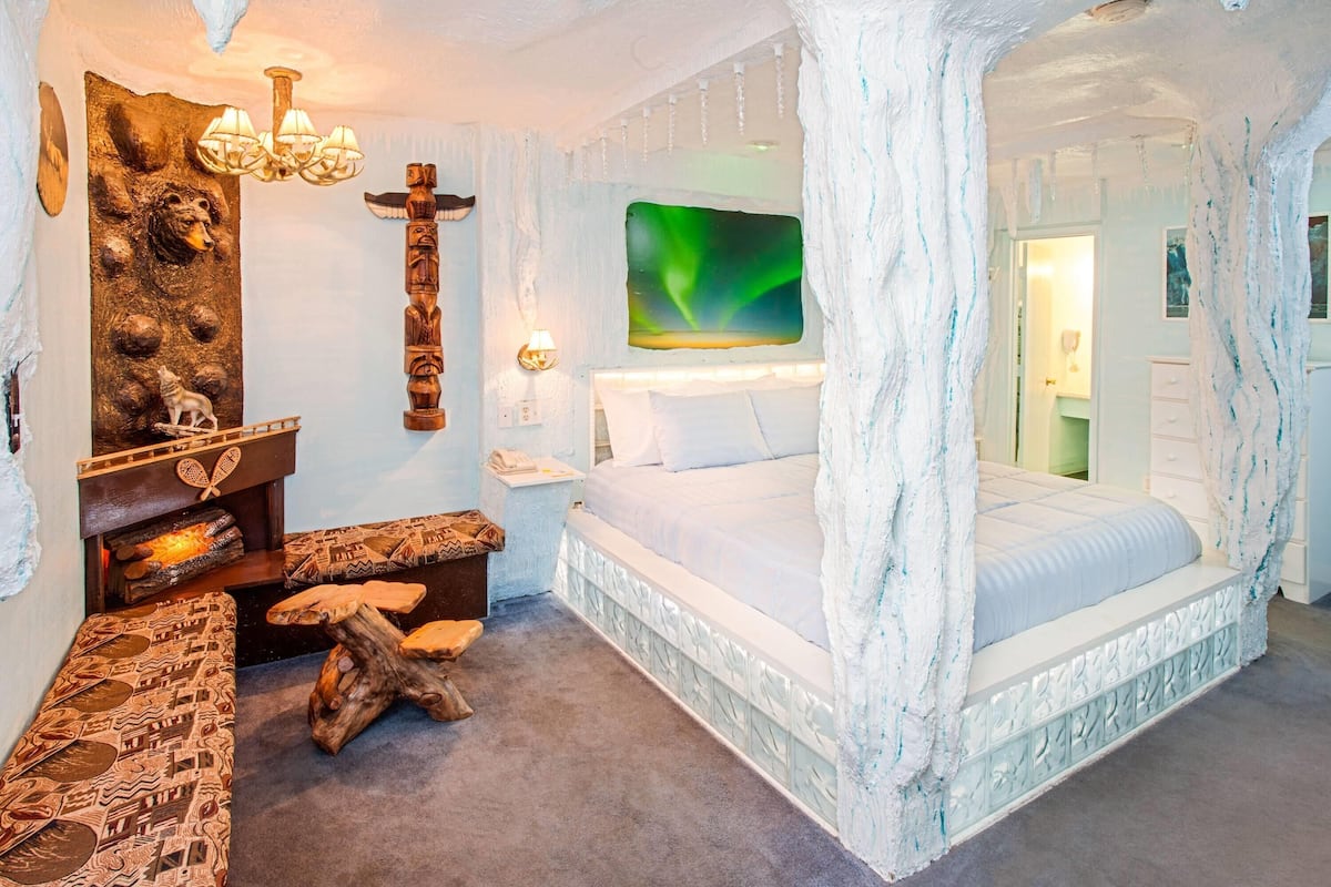 A room with a bed, a wooden bench, a wall with carvings, a chandelier, and a painting of the aurora borealis.
