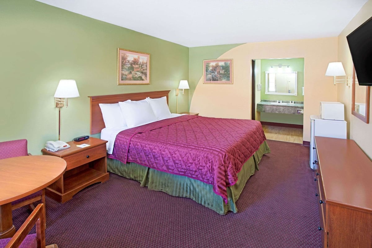 A hotel room with a bed, bedside tables, a desk, a chair, a television, and a bathroom visible through an open door.