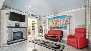 Una sala de estar con chimenea, un sofá rojo, un sillón rojo, un televisor de pantalla plana y un cuadro grande de un palacio.