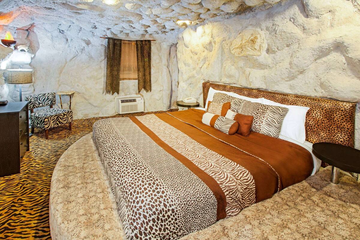A room with a stone wall, a bed with a zebra print bedspread, a zebra print chair, and a small table with a lamp.