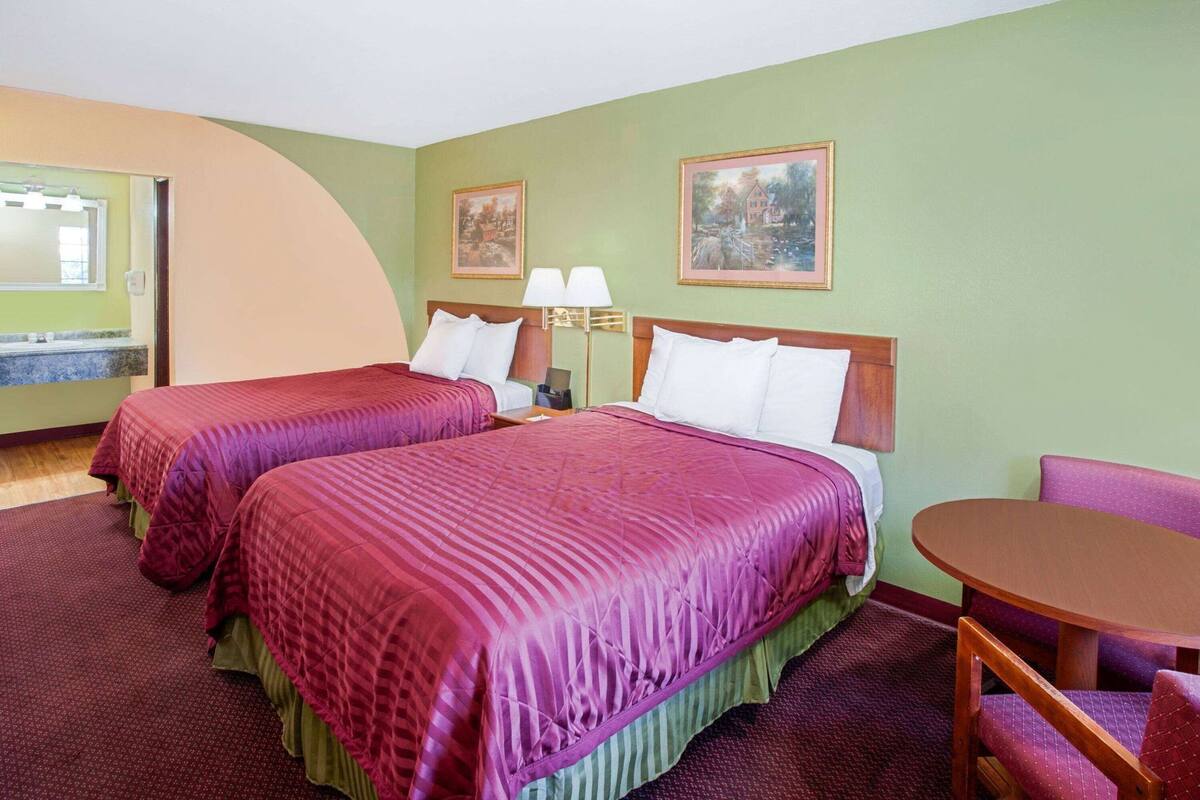 A hotel room with two beds, a small table, a chair, a lamp, and two framed pictures on the wall.