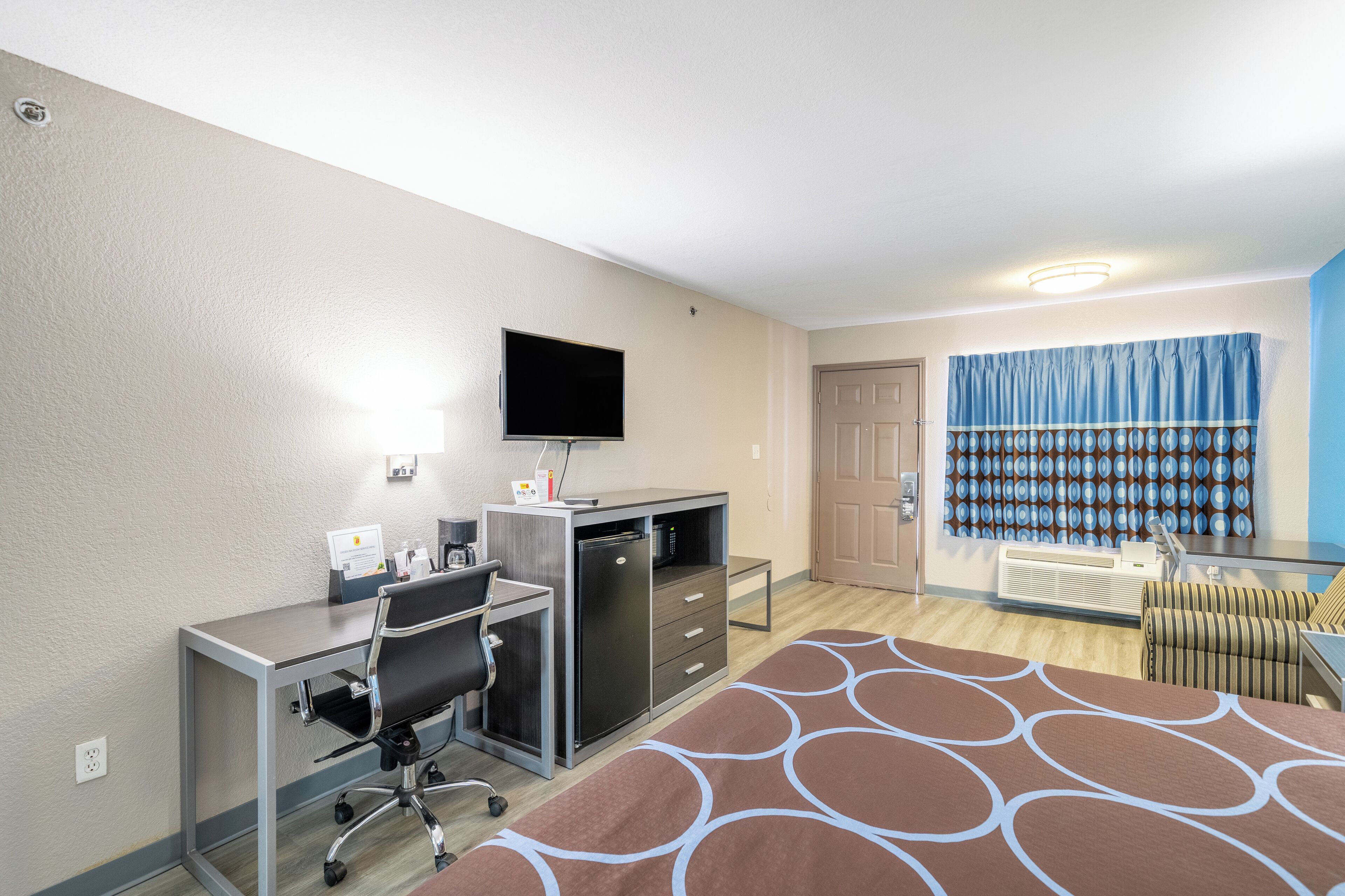 Une chambre d’hôtel comprenant un lit, un bureau avec une chaise, une télévision, un minibar et une fenêtre avec des rideaux.