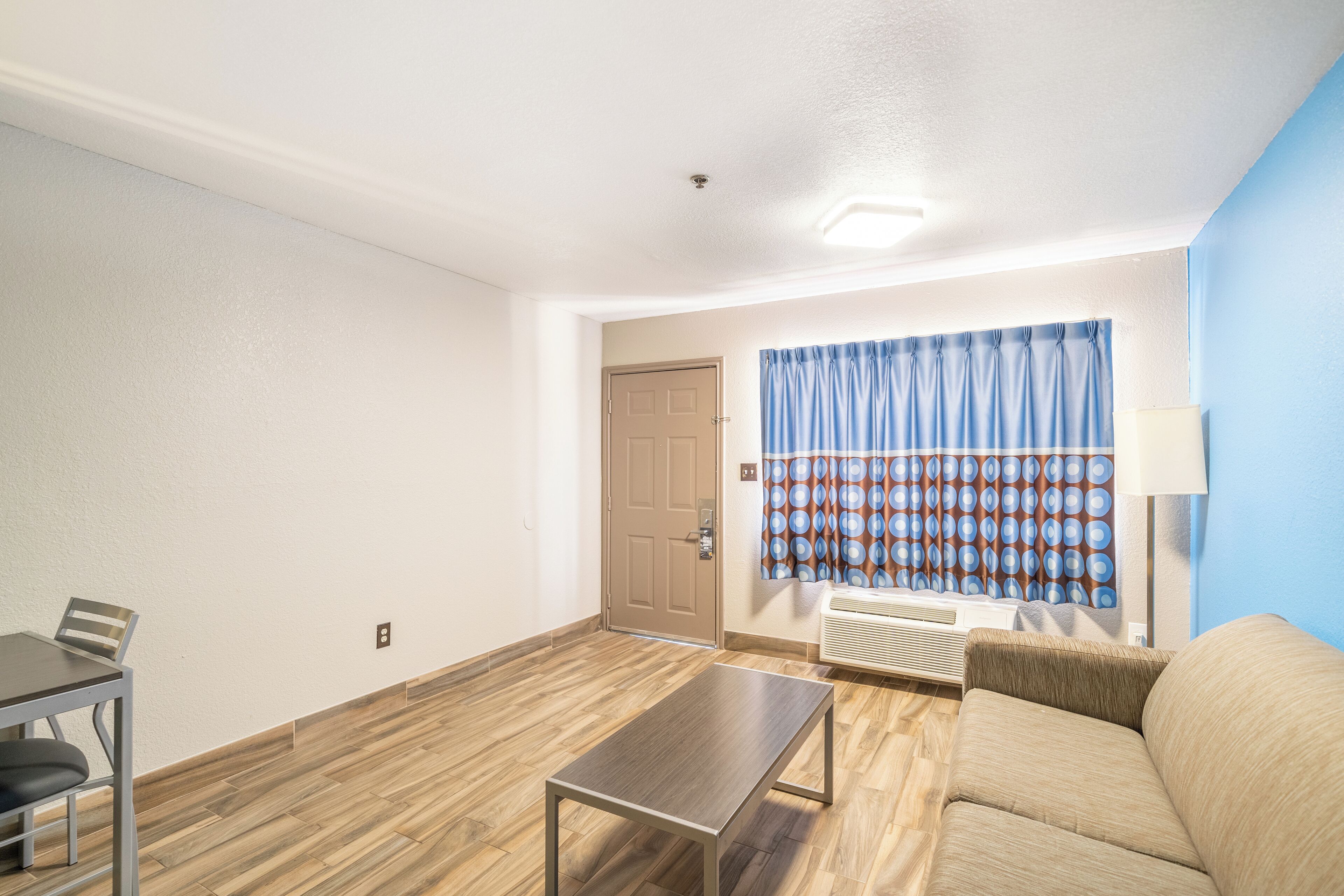 A room with a wooden floor, a beige sofa, a small table, a chair, a door, and a window with curtains.