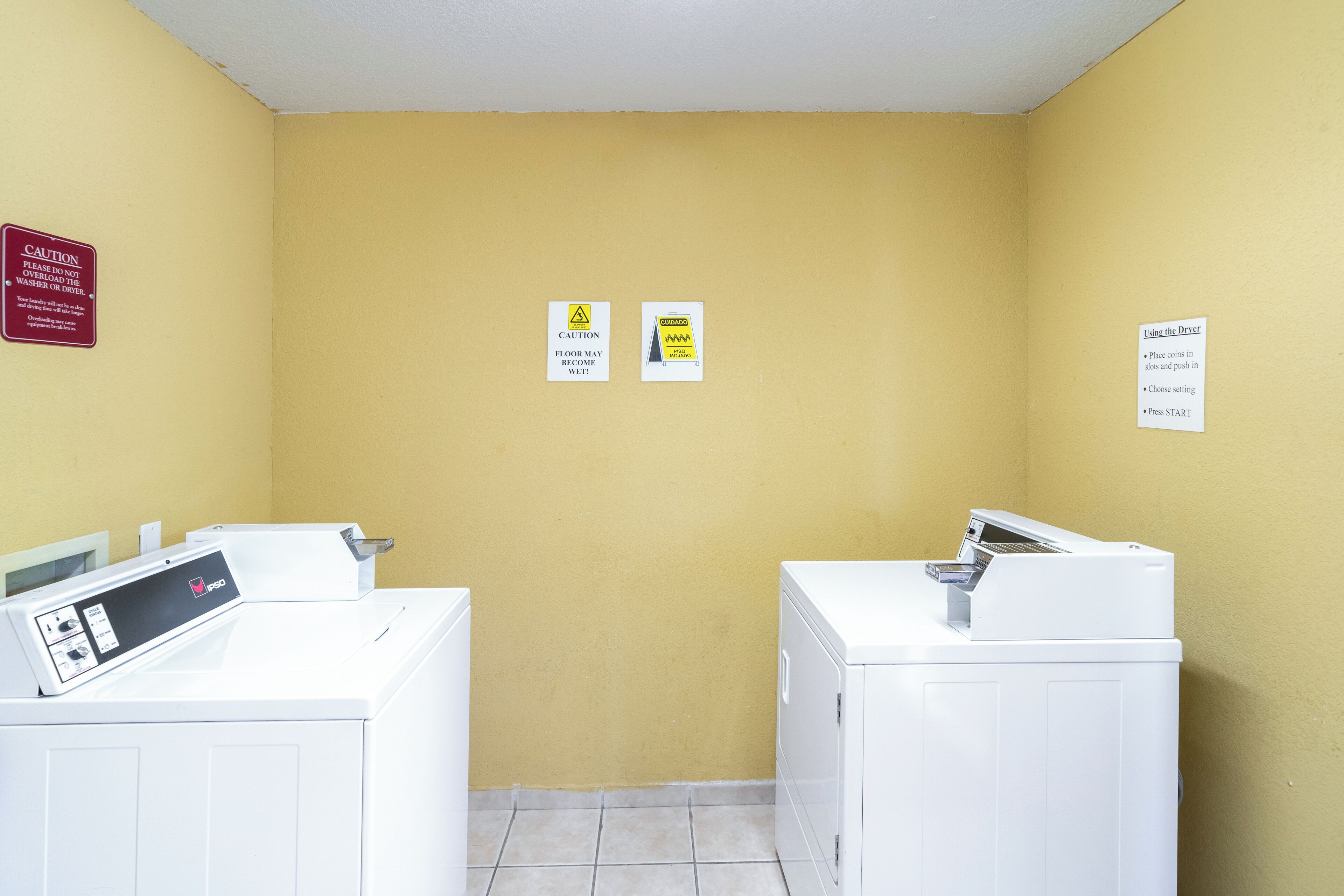 A laundry room with two washing machines, caution signs on the wall, and a floor with white tiles.