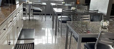 Interior de restaurante con zona de cocina, mesas, sillas y un amplio ventanal.